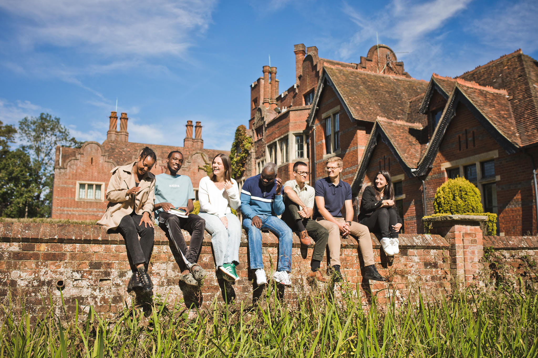 A multicultural group of Newbold students sitting along a sunlit wall, talking and laughing together
